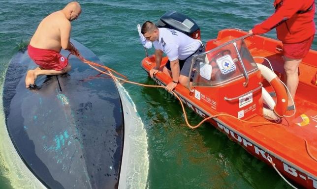 Dwie osoby w wodzie bez założonych kamizelek i łódź odwrócona do góry dnem